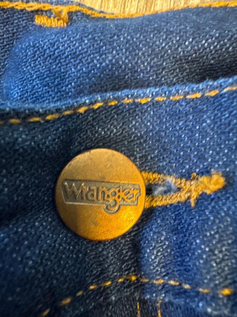 May include: Close-up of a blue denim jean with a gold Wrangler button.