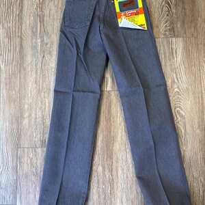 May include: A pair of gray Wrangler ProRodeo Cowboy Cut jeans. The jeans have a classic five-pocket design, a straight leg, and a Wrangler tag on the back pocket. The jeans are displayed on a wooden surface.