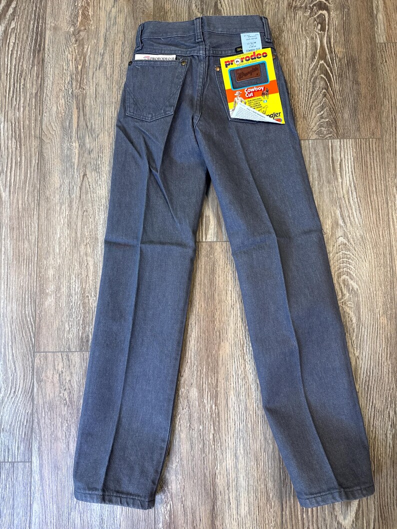 May include: A pair of gray Wrangler ProRodeo Cowboy Cut jeans. The jeans have a classic five-pocket design, a straight leg, and a Wrangler tag on the back pocket. The jeans are displayed on a wooden surface.