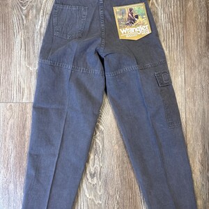 K&ouml;nnte beinhalten: Dunkelgraue Wrangler Rugged Wear Hose mit mehreren Taschen und einem R&uuml;ckenaufn&auml;her. Die Hose hat ein gerades Bein-Design und ein Etikett mit einer Grafik einer Person und eines Hundes.