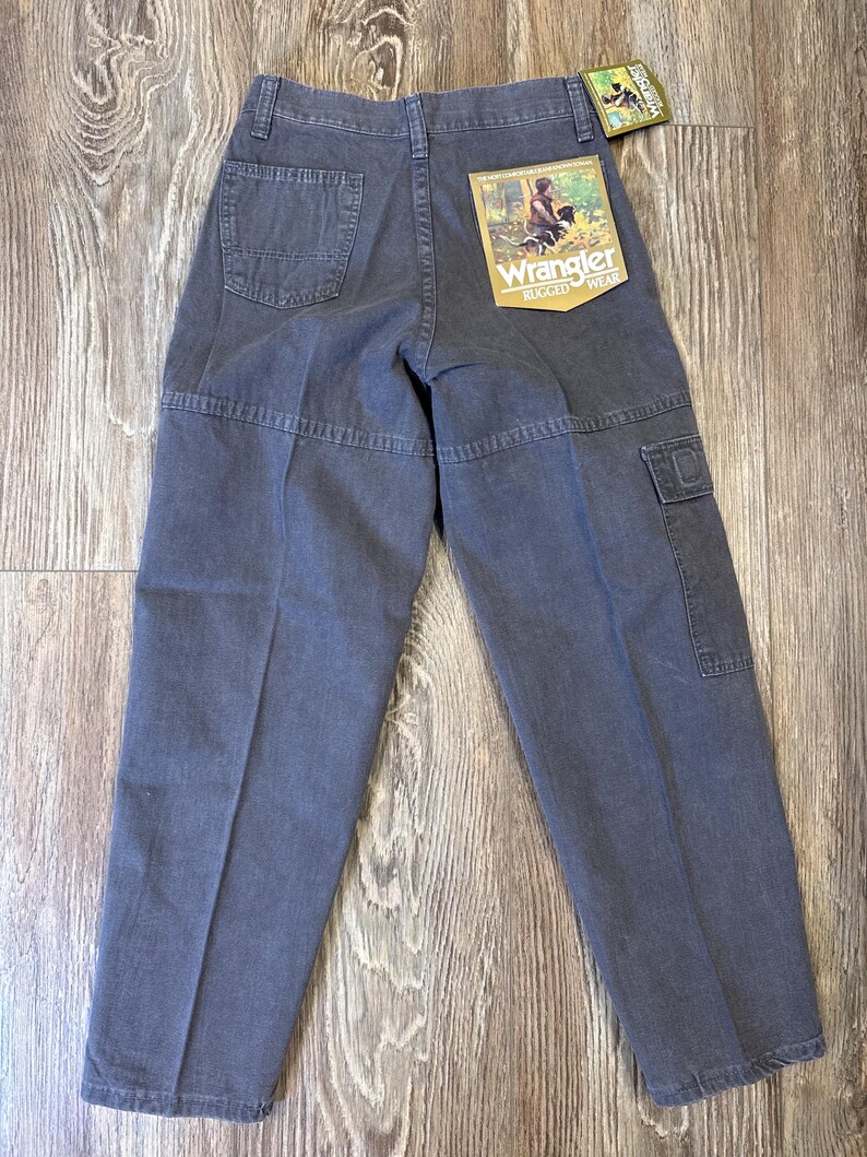 K&ouml;nnte beinhalten: Dunkelgraue Wrangler Rugged Wear Hose mit mehreren Taschen und einem R&uuml;ckenaufn&auml;her. Die Hose hat ein gerades Bein-Design und ein Etikett mit einer Grafik einer Person und eines Hundes.
