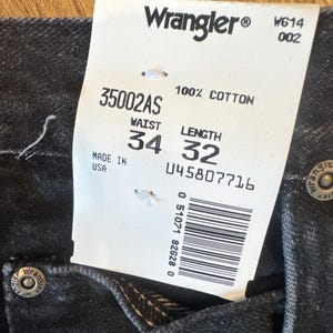 Puede incluir: Una etiqueta blanca con texto negro que dice "Wrangler&reg; W614 002 35002AS 100% ALGOD&Oacute;N CINTURA LARGO HECHO EN EE. UU. 34 32 045807716 0 51071 82628 0" con un c&oacute;digo de barras. La etiqueta est&aacute; unida a un par de pantalones vaqueros negros.