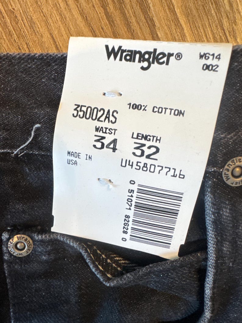 Puede incluir: Una etiqueta blanca con texto negro que dice "Wrangler&reg; W614 002 35002AS 100% ALGOD&Oacute;N CINTURA LARGO HECHO EN EE. UU. 34 32 045807716 0 51071 82628 0" con un c&oacute;digo de barras. La etiqueta est&aacute; unida a un par de pantalones vaqueros negros.