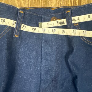 K&ouml;nnte beinhalten: Nahaufnahme des Bundes einer dunkelblauen Jeans. Ein Ma&szlig;band zeigt eine Bundweite von ca. 74 cm. Die Jeans hat einen Knopfverschluss und orangefarbene N&auml;hte.