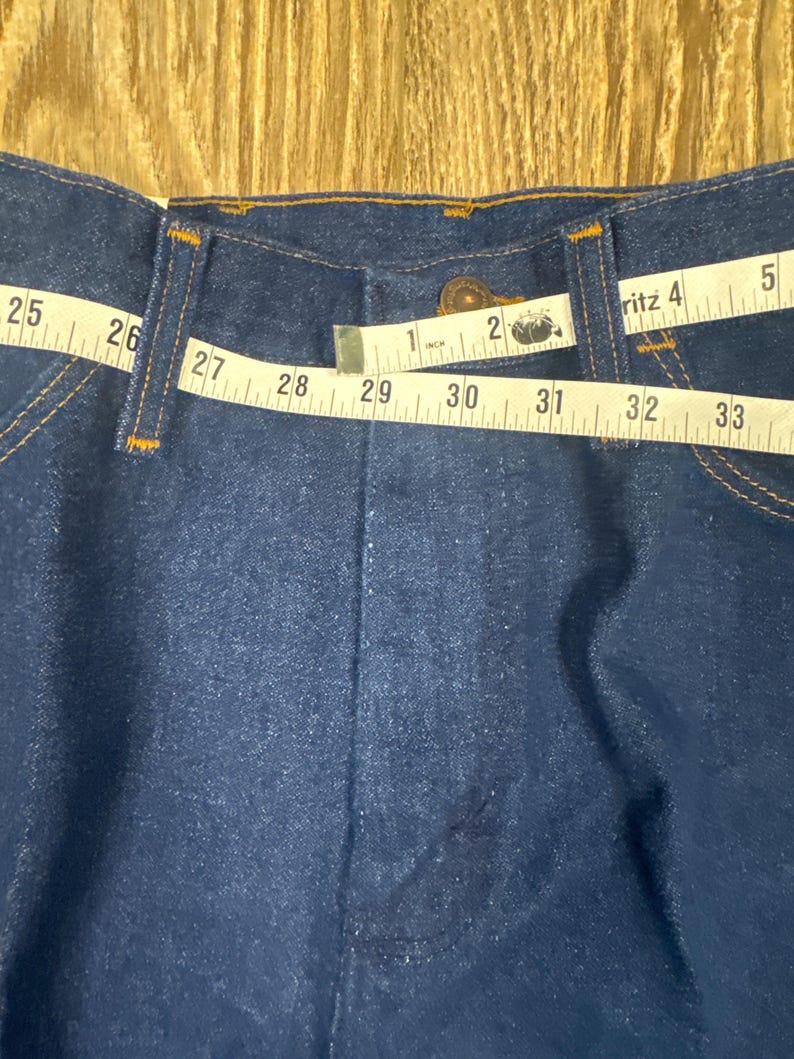 K&ouml;nnte beinhalten: Nahaufnahme des Bundes einer dunkelblauen Jeans. Ein Ma&szlig;band zeigt eine Bundweite von ca. 74 cm. Die Jeans hat einen Knopfverschluss und orangefarbene N&auml;hte.