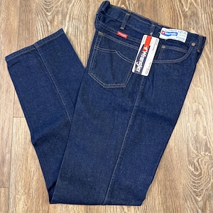May include: Dark blue Wrangler jeans with a straight leg design. The jeans have a classic five-pocket style, a zipper fly, and a Wrangler tag. The jeans are folded and laid on a wooden surface.