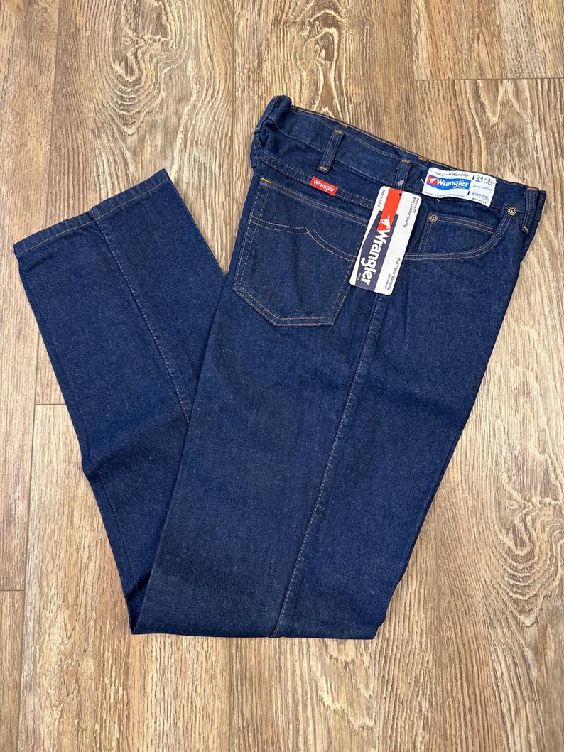 May include: Dark blue Wrangler jeans with a straight leg design. The jeans have a classic five-pocket style, a zipper fly, and a Wrangler tag. The jeans are folded and laid on a wooden surface.