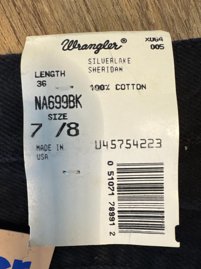 K&ouml;nnte beinhalten: Ein wei&szlig;es Etikett mit schwarzem Text, das auf eine Wrangler Silverlake Sheridan Jeans hinweist. Das Etikett zeigt die Gr&ouml;&szlig;e 7/8, die L&auml;nge 36, den Stil NA699BK und den Stoff 100% Baumwolle. Das Etikett enth&auml;lt auch einen Barcode und den Text "Hergestellt in den USA".