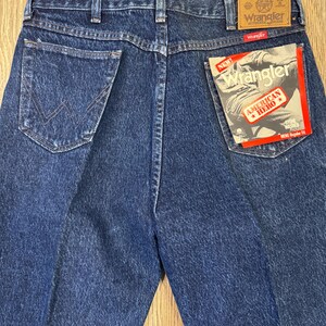 May include: Back view of blue denim Wrangler jeans. The jeans feature a classic five-pocket design, with the Wrangler label on the back pocket. A tag reads "New Wrangler American Hero".
