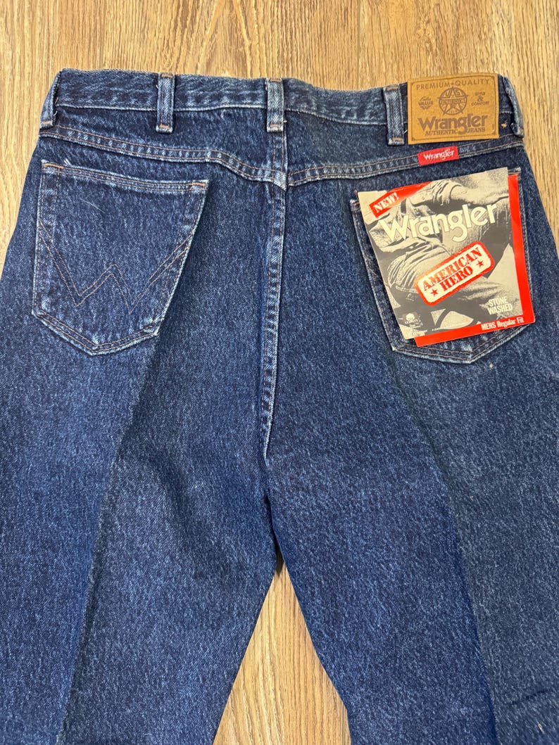 May include: Back view of blue denim Wrangler jeans. The jeans feature a classic five-pocket design, with the Wrangler label on the back pocket. A tag reads "New Wrangler American Hero".