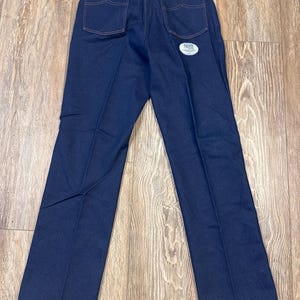May include: Dark blue denim jeans with orange stitching. The jeans have two back pockets and a small red Wrangler label. A white tag is attached to the jeans.