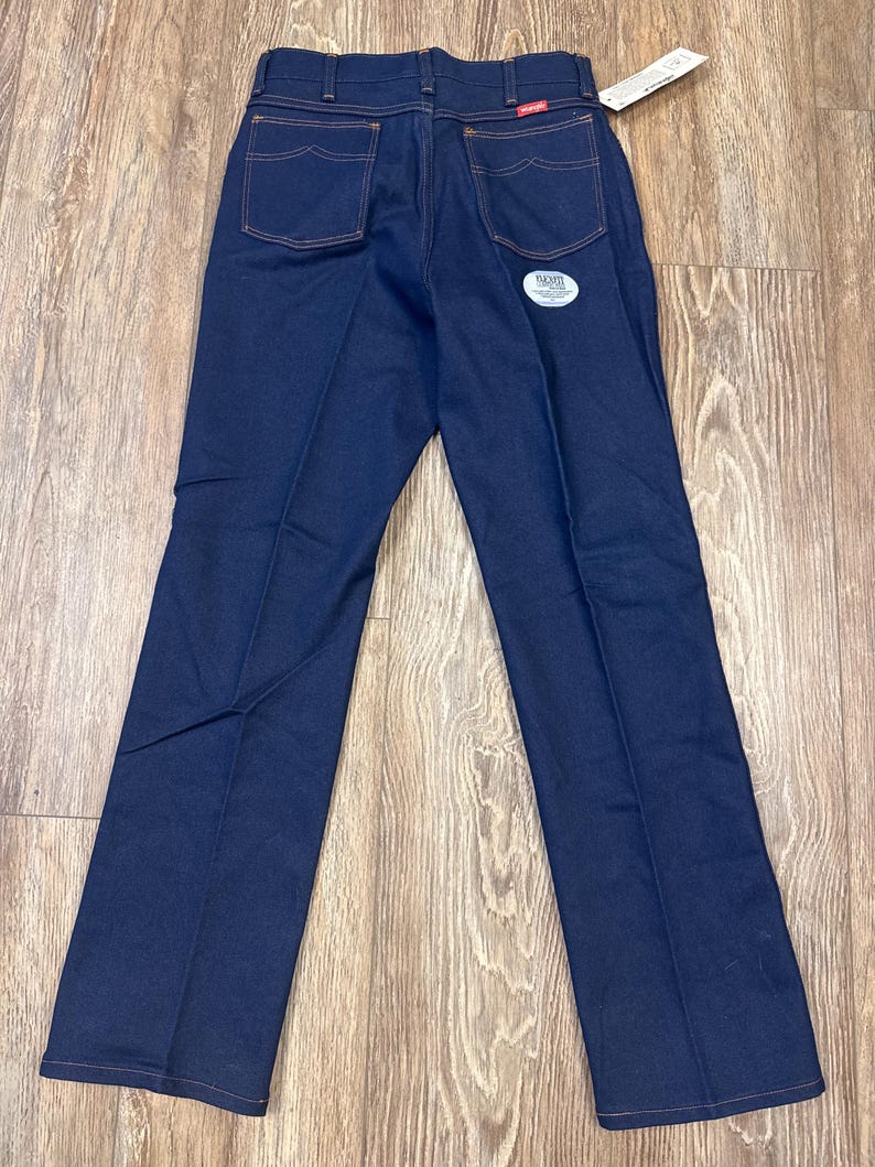 May include: Dark blue denim jeans with orange stitching. The jeans have two back pockets and a small red Wrangler label. A white tag is attached to the jeans.