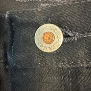 May include: Close-up of a black denim garment with a silver button. The button has the words "Silver Lake" around the edge and a copper-colored center. The fabric has a textured appearance.