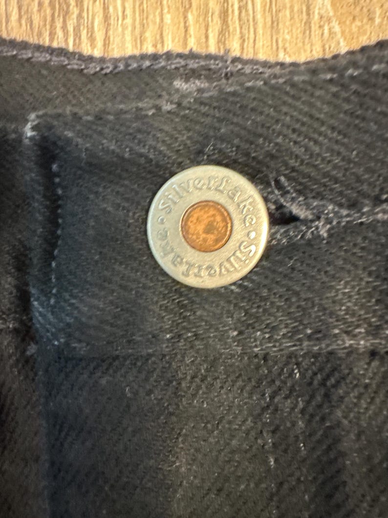 May include: Close-up of a black denim garment with a silver button. The button has the words "Silver Lake" around the edge and a copper-colored center. The fabric has a textured appearance.