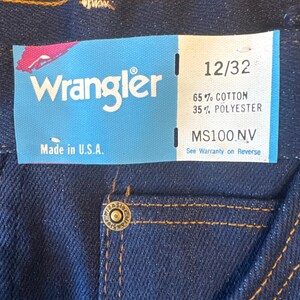 K&ouml;nnte beinhalten: Eine blaue Jeans aus Denim mit einem braunen Leder-Patch und einem Wrangler-Etikett. Das Etikett zeigt "Wrangler" und "Made in U.S.A.". Das Gr&ouml;&szlig;enetikett zeigt "12/32", "65% Baumwolle 35% Polyester" und "MS100.NV".