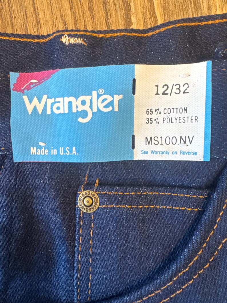 K&ouml;nnte beinhalten: Eine blaue Jeans aus Denim mit einem braunen Leder-Patch und einem Wrangler-Etikett. Das Etikett zeigt "Wrangler" und "Made in U.S.A.". Das Gr&ouml;&szlig;enetikett zeigt "12/32", "65% Baumwolle 35% Polyester" und "MS100.NV".