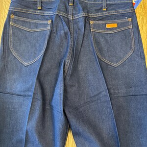May include: A pair of dark blue denim jeans with a brown leather patch on the back pocket. The jeans have a classic straight leg fit and are made by Wrangler.