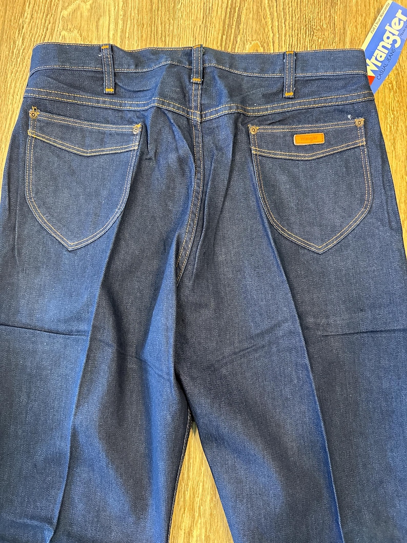 May include: A pair of dark blue denim jeans with a brown leather patch on the back pocket. The jeans have a classic straight leg fit and are made by Wrangler.