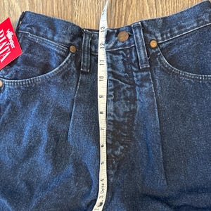 May include: Close-up of dark blue denim jeans with a high waist. A measuring tape shows the waist is approximately 13 inches. The jeans have classic five-pocket styling with copper-colored rivets and a Wrangler tag.