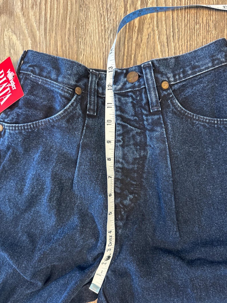 May include: Close-up of dark blue denim jeans with a high waist. A measuring tape shows the waist is approximately 13 inches. The jeans have classic five-pocket styling with copper-colored rivets and a Wrangler tag.