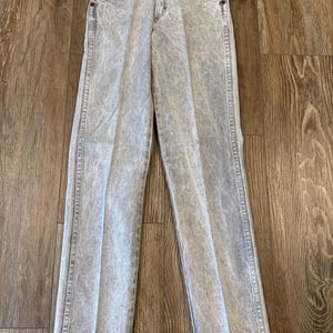 May include: Acid wash gray denim jeans with a classic five-pocket design. The jeans have a straight leg cut and a button and zipper closure. A small tag is attached to the jeans.