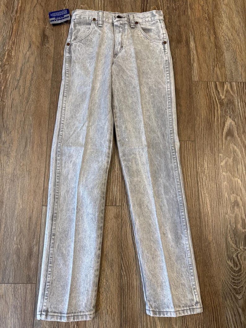 May include: Acid wash gray denim jeans with a classic five-pocket design. The jeans have a straight leg cut and a button and zipper closure. A small tag is attached to the jeans.