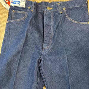 May include: Close-up of a pair of dark blue Wrangler jeans. The jeans have a classic five-pocket design with a button and zipper closure. The tag reads "Wrangler" and indicates a 34x32 size. The stitching is a contrasting tan color.