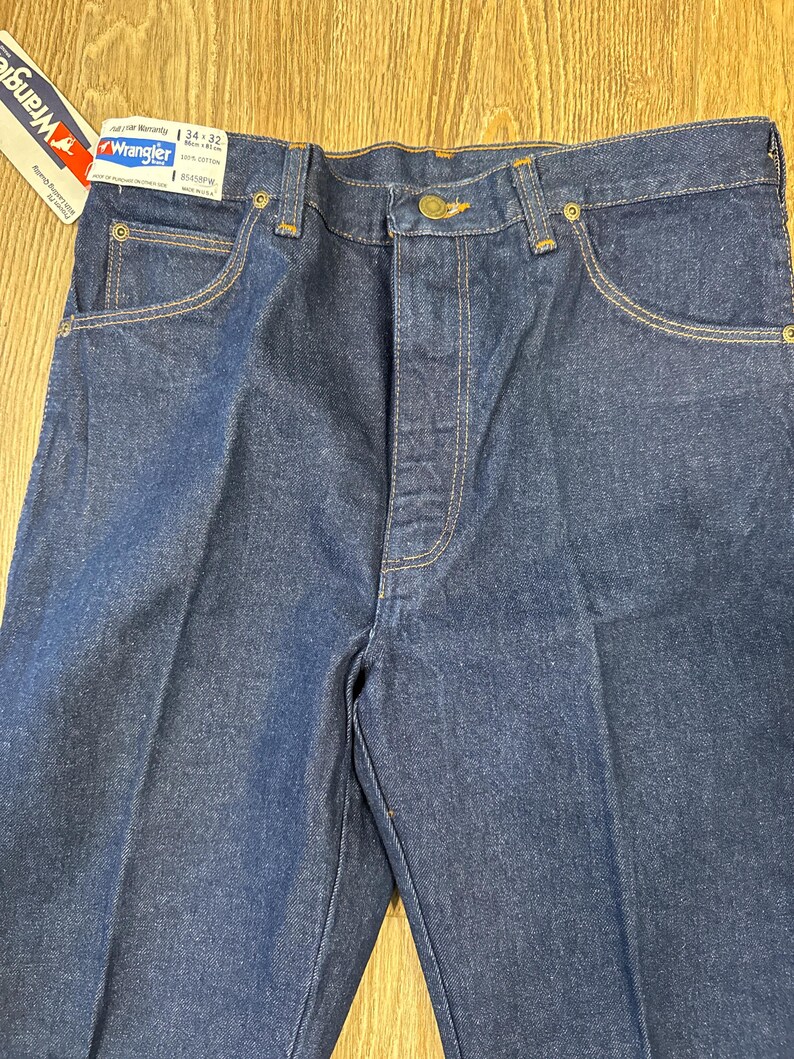 May include: Close-up of a pair of dark blue Wrangler jeans. The jeans have a classic five-pocket design with a button and zipper closure. The tag reads "Wrangler" and indicates a 34x32 size. The stitching is a contrasting tan color.