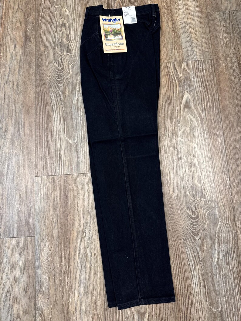 May include: Dark wash Wrangler Silverlake jeans. The jeans are folded and have a tag attached with the Wrangler logo and product information. The jeans are made of a dark blue denim material and are laid out on a light brown wooden surface.