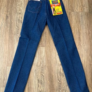 May include: A pair of blue denim jeans with a yellow Wrangler ProRodeo label attached to the back waistband. The label features a cowboy riding a horse and the text "Cowboy Cut".