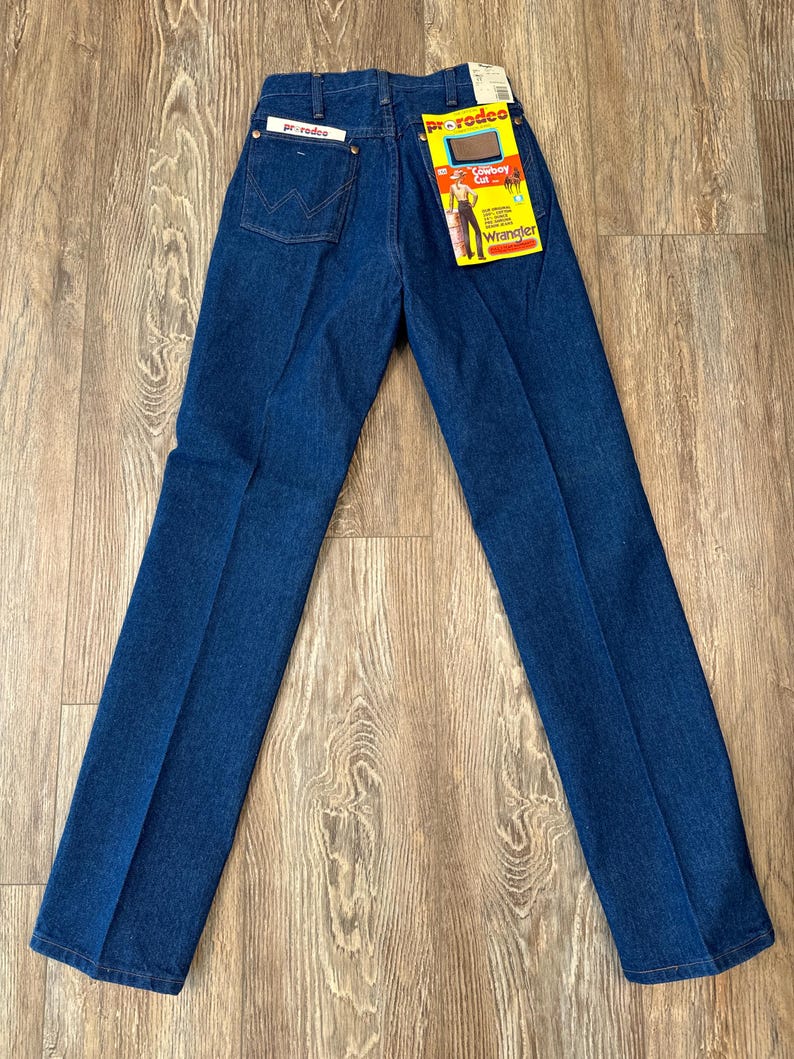 May include: A pair of blue denim jeans with a yellow Wrangler ProRodeo label attached to the back waistband. The label features a cowboy riding a horse and the text "Cowboy Cut".