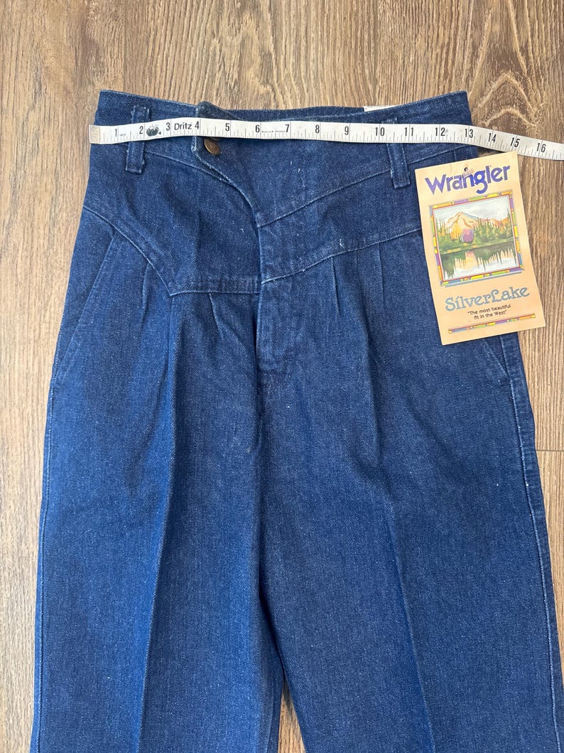 Puede incluir: Vaqueros Wrangler Silverlake de mezclilla azul con cintura alta y pliegues. Una cinta m&eacute;trica est&aacute; colocada sobre la cintura. La etiqueta dice "Wrangler SilverLake".