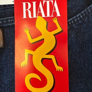 May include: A red tag with the Wrangler Riata logo and a yellow lizard graphic. The tag reads "WOMEN'S WESTERN RELAXED FIT". The tag is attached to a pair of blue denim jeans.