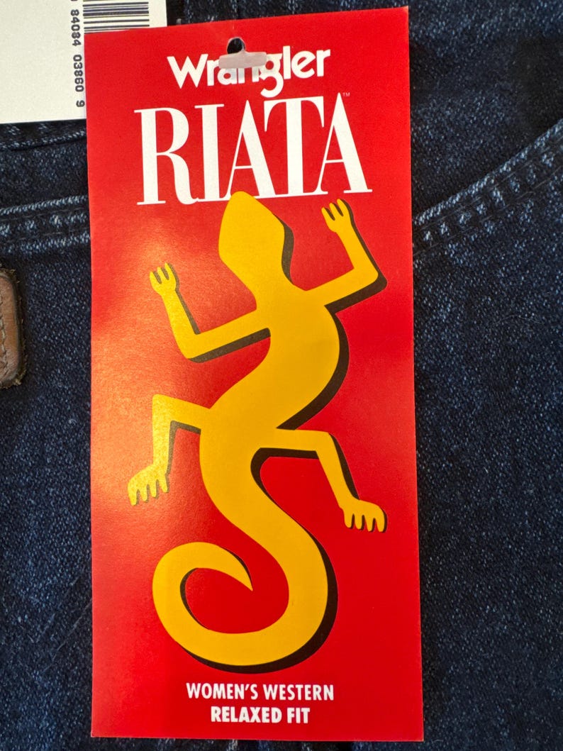 May include: A red tag with the Wrangler Riata logo and a yellow lizard graphic. The tag reads "WOMEN'S WESTERN RELAXED FIT". The tag is attached to a pair of blue denim jeans.