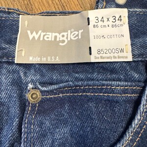 K&ouml;nnte beinhalten: Nahaufnahme einer blauen Wrangler-Jeans. Das Etikett lautet "34 x 34" und "86 cm x 86 cm", mit "100% COTTON" und "Made in U.S.A." aufgedruckt. Die Jeans hat einen Knopf und eine Tasche.