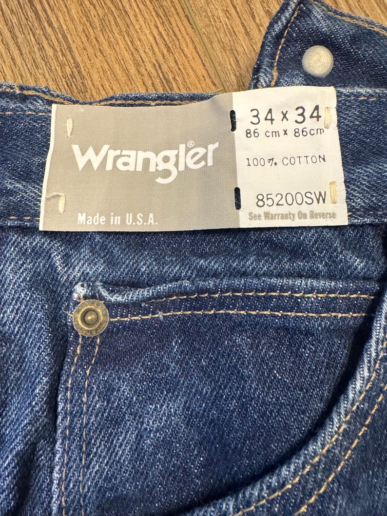 K&ouml;nnte beinhalten: Nahaufnahme einer blauen Wrangler-Jeans. Das Etikett lautet "34 x 34" und "86 cm x 86 cm", mit "100% COTTON" und "Made in U.S.A." aufgedruckt. Die Jeans hat einen Knopf und eine Tasche.