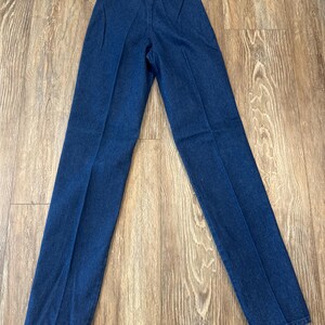 Puede incluir: Pantalones vaqueros azul oscuro con cintura alta y pernera recta. Los pantalones tienen una etiqueta Wrangler adjunta. Los pantalones est&aacute;n colocados sobre una superficie de madera.