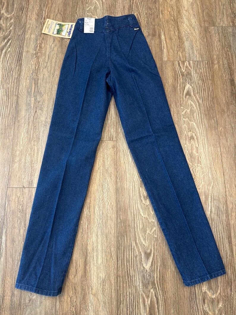 Puede incluir: Pantalones vaqueros azul oscuro con cintura alta y pernera recta. Los pantalones tienen una etiqueta Wrangler adjunta. Los pantalones est&aacute;n colocados sobre una superficie de madera.