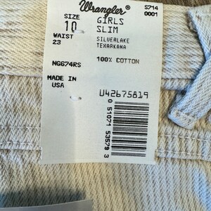 May include: A white Wrangler girls' slim jeans tag with the size 10 and waist 23. The tag also says "Silverlake Texarkana" and "100% Cotton". The tag has a barcode and the text "NG674RS" and "Made in USA".