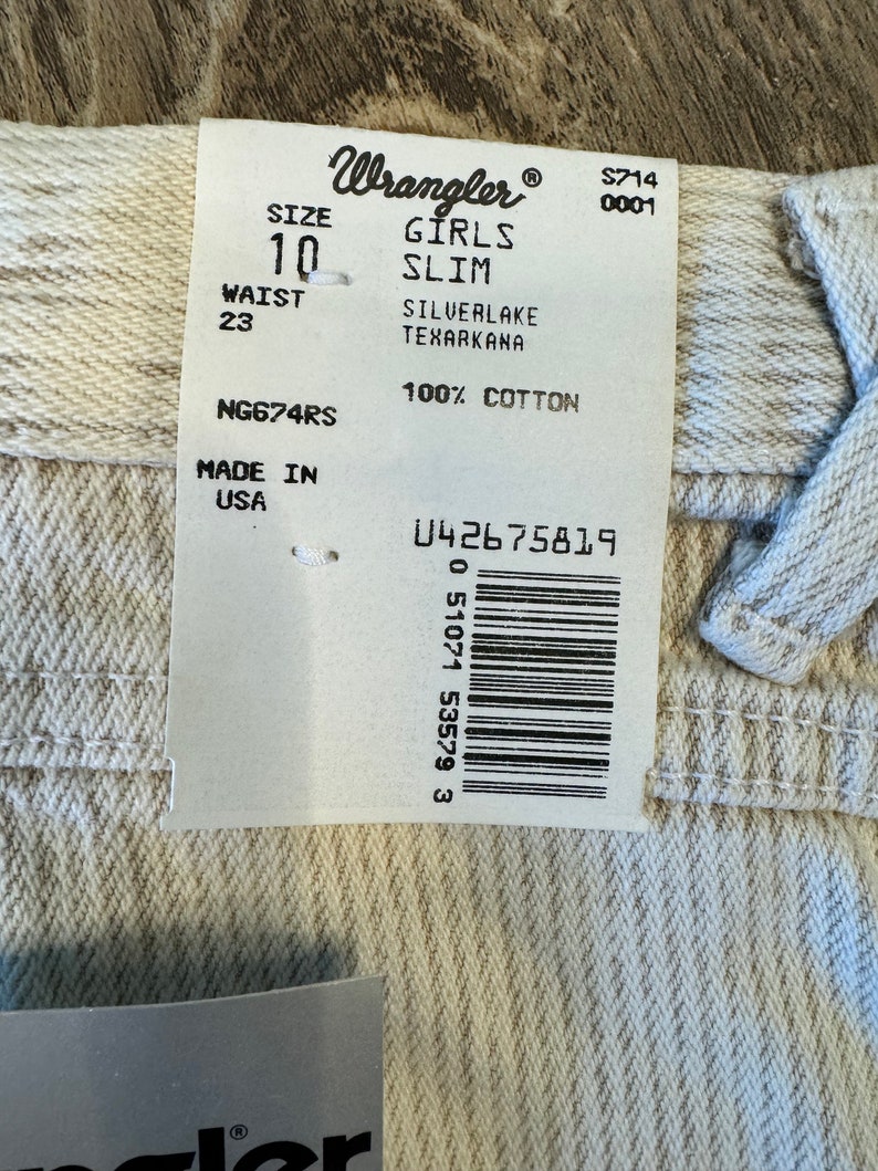 May include: A white Wrangler girls' slim jeans tag with the size 10 and waist 23. The tag also says "Silverlake Texarkana" and "100% Cotton". The tag has a barcode and the text "NG674RS" and "Made in USA".
