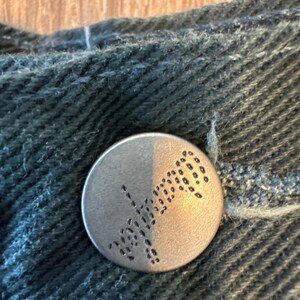 May include: A silver button with a black design of a stylized flower or leaf. The button is on a dark blue denim fabric.