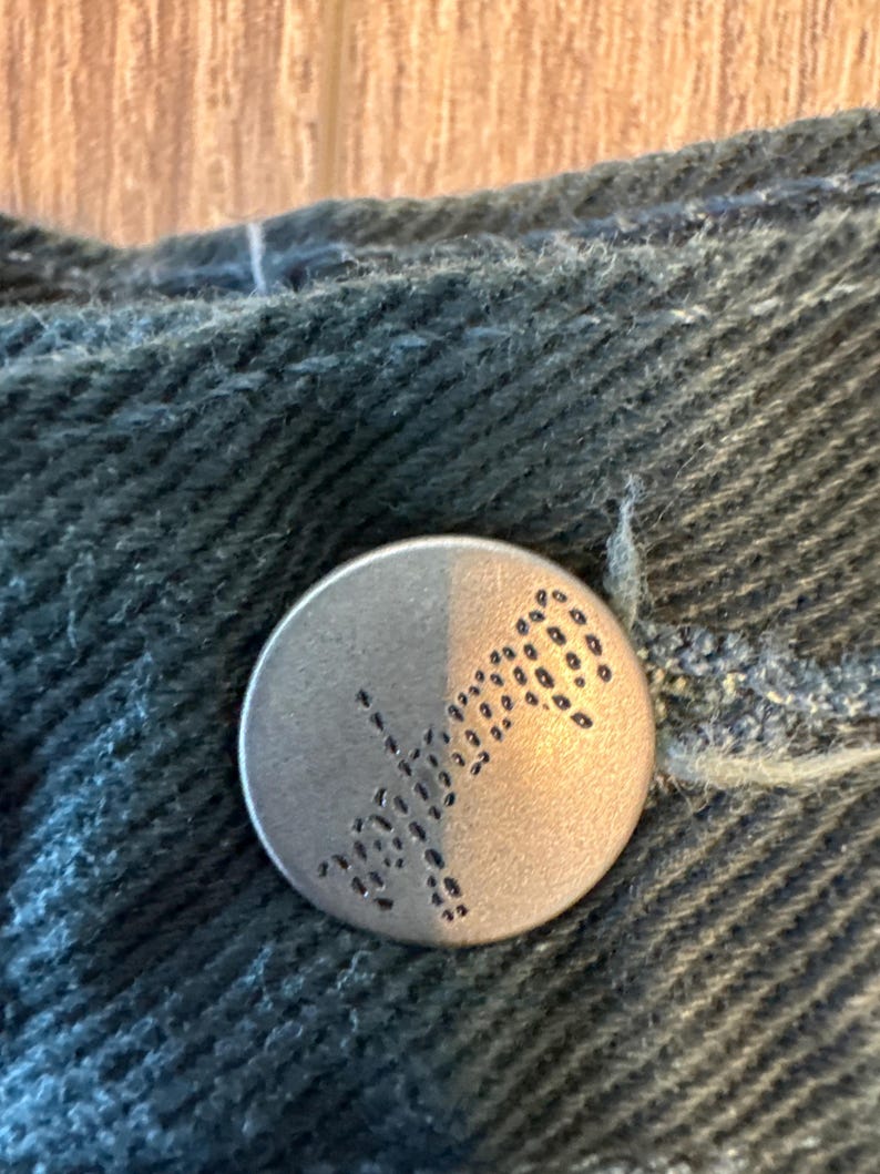 May include: A silver button with a black design of a stylized flower or leaf. The button is on a dark blue denim fabric.