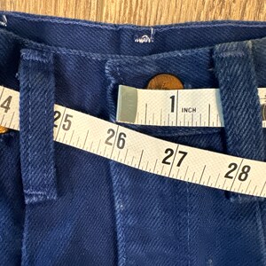 May include: A close-up of a dark blue pair of jeans with a measuring tape across the waistband. The tape reads 25, 26, 27, 28, and 1 inch.