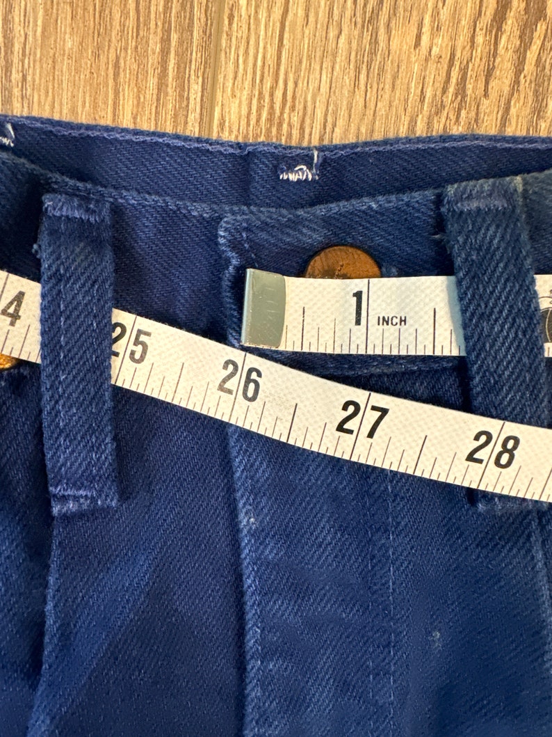 May include: A close-up of a dark blue pair of jeans with a measuring tape across the waistband. The tape reads 25, 26, 27, 28, and 1 inch.