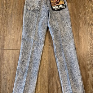 K&ouml;nnte beinhalten: Hellblaue Jeans von Wrangler Rugged Wear mit S&auml;urewaschung. Die Jeans haben ein klassisches F&uuml;nf-Taschen-Design, ein gerades Bein und einen Wrangler-Patch auf der Ges&auml;&szlig;tasche. Ein Anh&auml;nger ist an der Jeans befestigt.