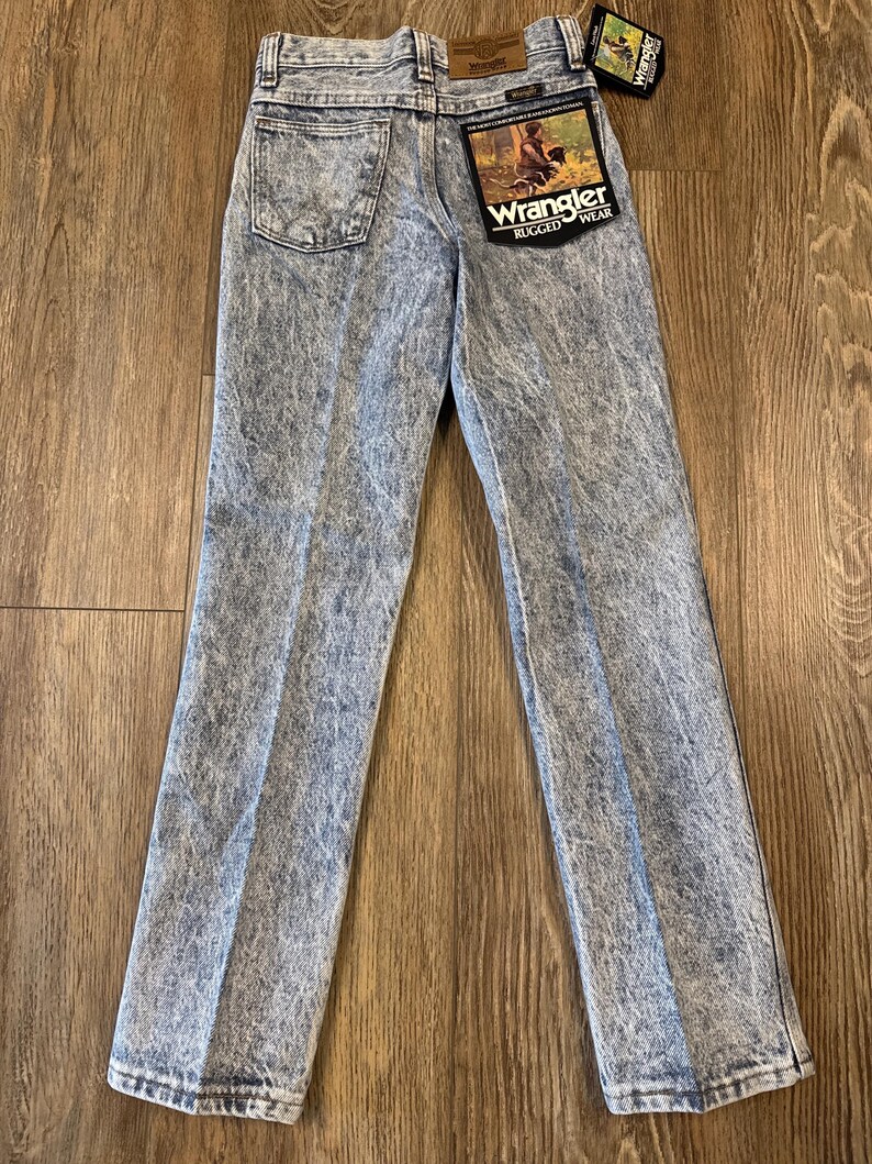 K&ouml;nnte beinhalten: Hellblaue Jeans von Wrangler Rugged Wear mit S&auml;urewaschung. Die Jeans haben ein klassisches F&uuml;nf-Taschen-Design, ein gerades Bein und einen Wrangler-Patch auf der Ges&auml;&szlig;tasche. Ein Anh&auml;nger ist an der Jeans befestigt.