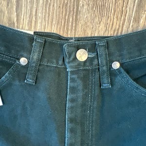 May include: Close-up of the waistband of a pair of dark green denim jeans. The jeans have a silver button and rivets.