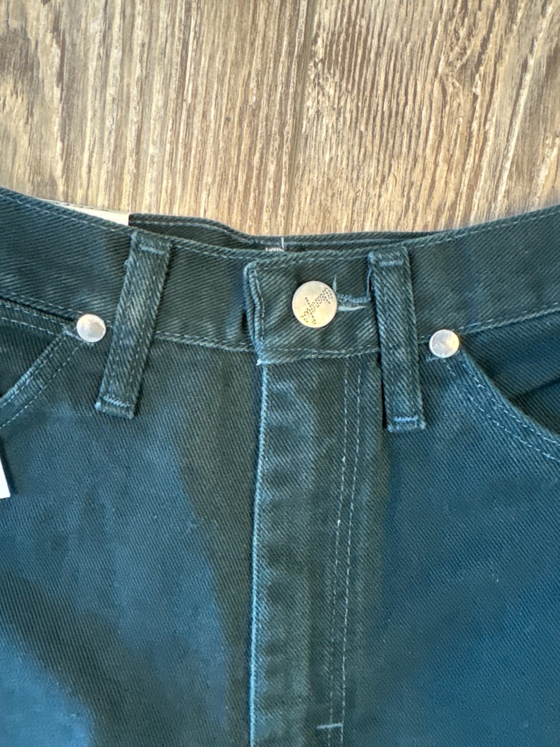 May include: Close-up of the waistband of a pair of dark green denim jeans. The jeans have a silver button and rivets.