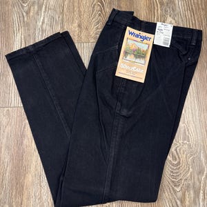 May include: Black Wrangler Silverlake jeans with a straight leg. The jeans have a classic five-pocket design and a zip fly. A tag with the Wrangler logo and "Silverlake" text is attached. The jeans are laid out on a wooden surface.