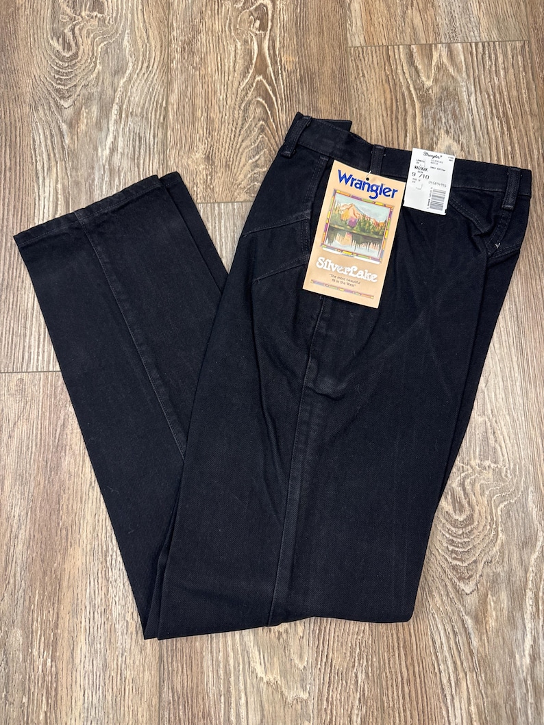 May include: Black Wrangler Silverlake jeans with a straight leg. The jeans have a classic five-pocket design and a zip fly. A tag with the Wrangler logo and "Silverlake" text is attached. The jeans are laid out on a wooden surface.